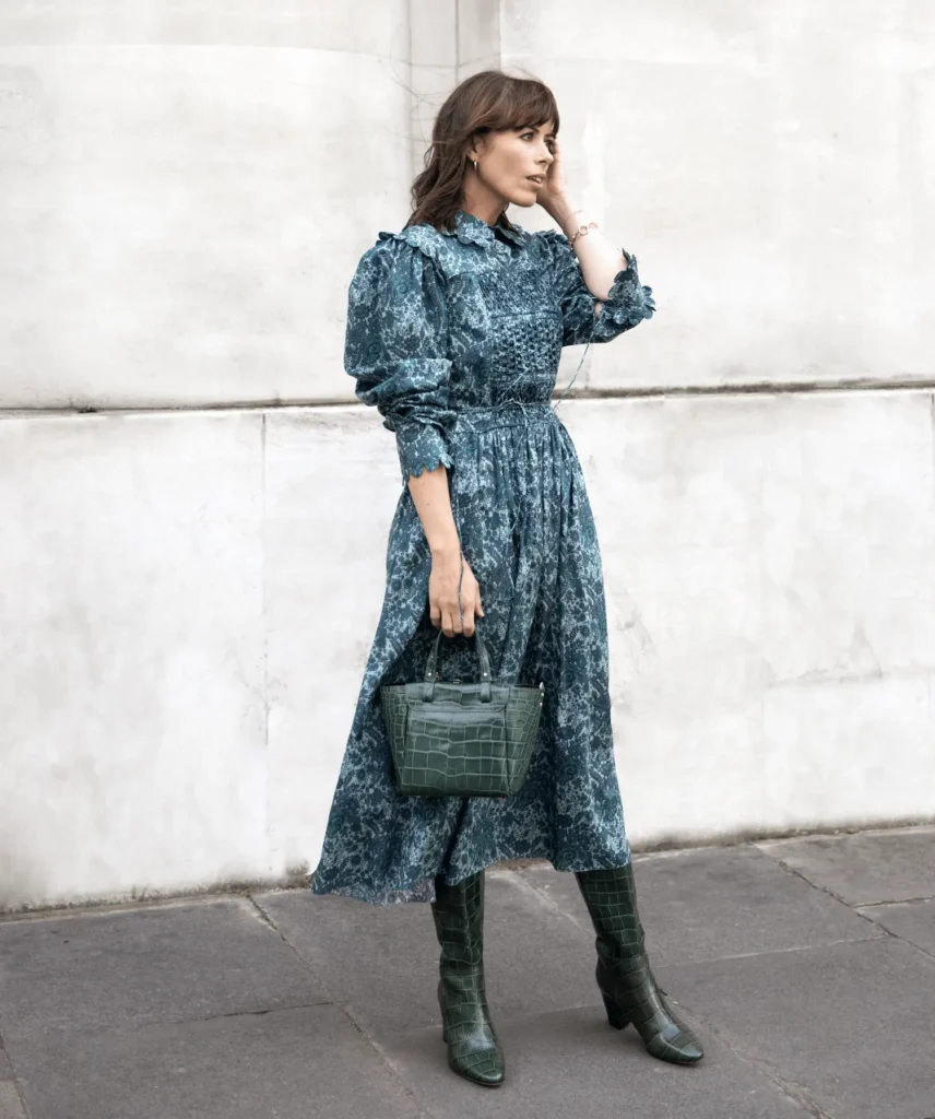 Woman in a teal patterned midi dress with puff sleeves, carrying a matching green crocodile-embossed handbag and wearing coordinating knee-high boots, standing against a light stone wall.