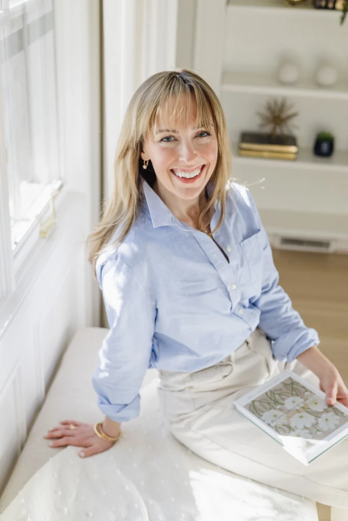 Portrait of Jen Shoop in a blue button-down shirt, holding her book, Small Wonders, by a sunlit window