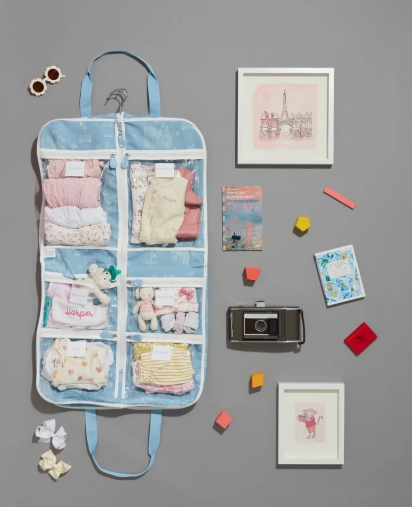 Flat lay of a blue hanging travel organizer packed with children’s clothes and accessories, styled with small books, framed prints, sunglasses, toy blocks, and a camera on a gray background.