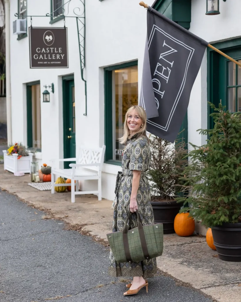 Jen Shoop standing outside Castle Gallery in Middleburg, beside its storefront sign and open flag.